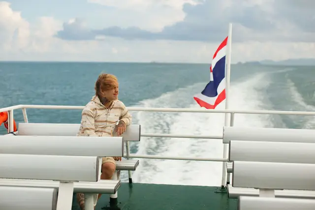 A passenger on the deck of a ferry, the Thai flag flying over the wake
