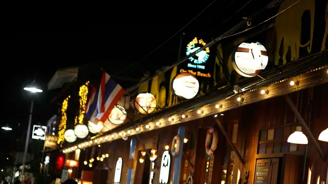 Enseignes lumineuses et drapeau thaïlandais devant un bar de bord de plage, la nuit