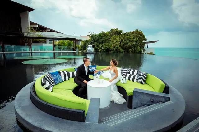 A newlywed couple in an outdoor lounge beside an infinity pool facing the sea